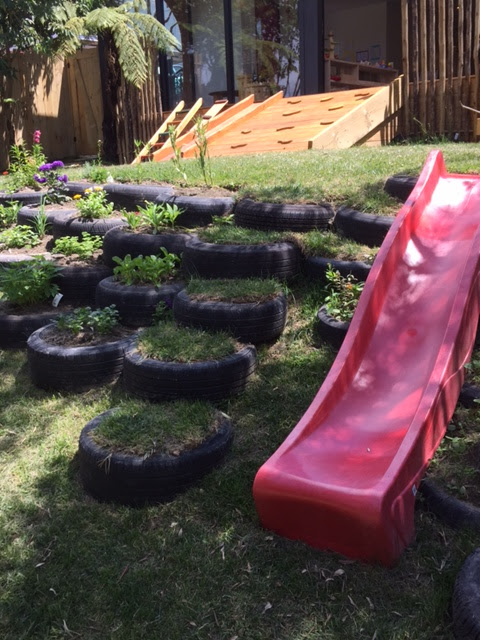 Red plastic slide placed on a low bank. Flowering plants are planted in recycled tires in the bank.
